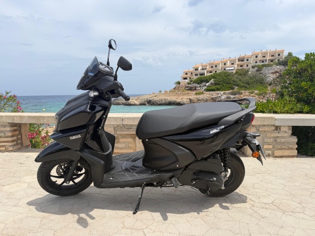Black scooter parked by seaside with buildings in background.